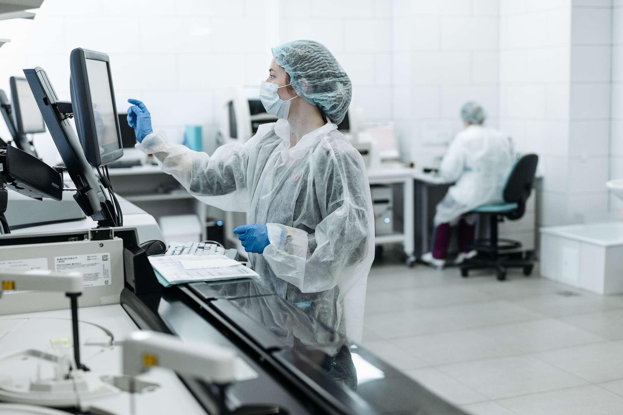 Kvinne i smittevernklær trykker på en skjerm i et laboratorium. Foto.