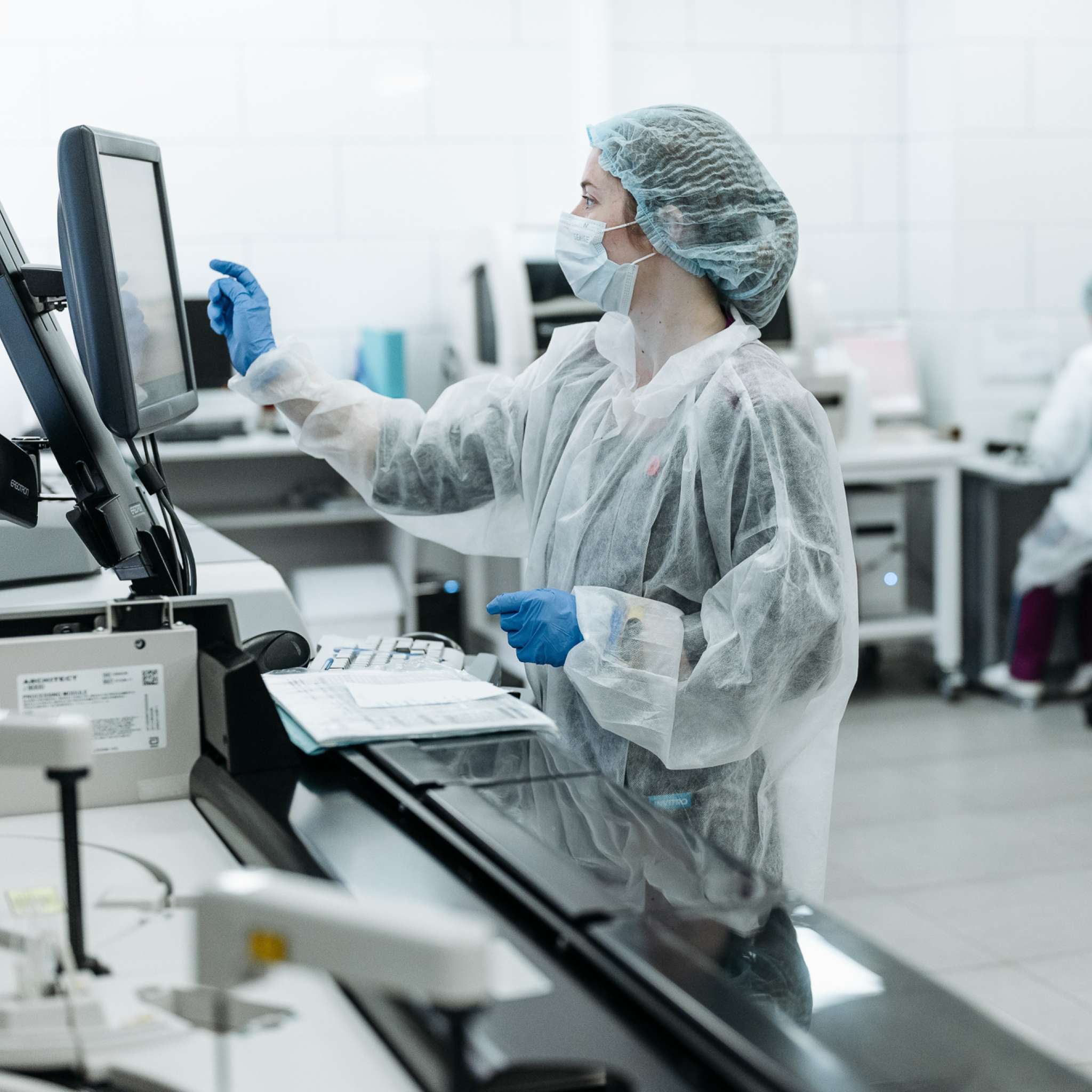 Kvinne i smittevernklær trykker på en skjerm i et laboratorium. Foto.